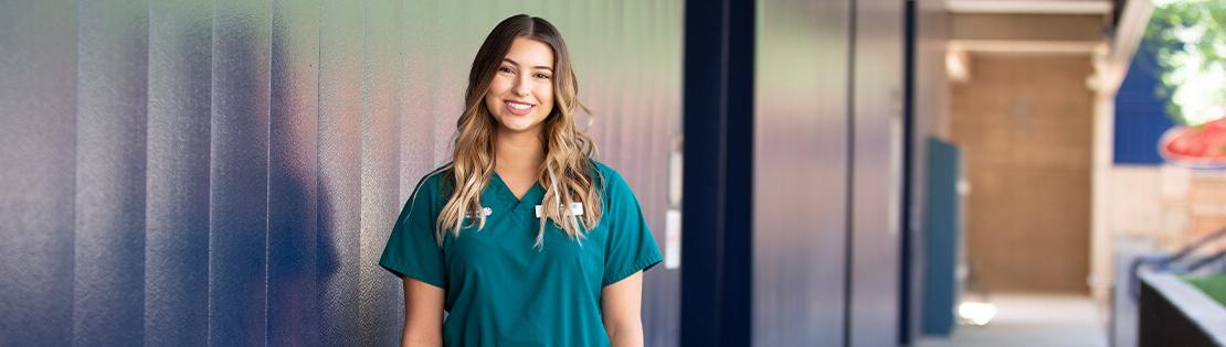 A Student in nursing scrubs stands smiling outside of Pima's West campus